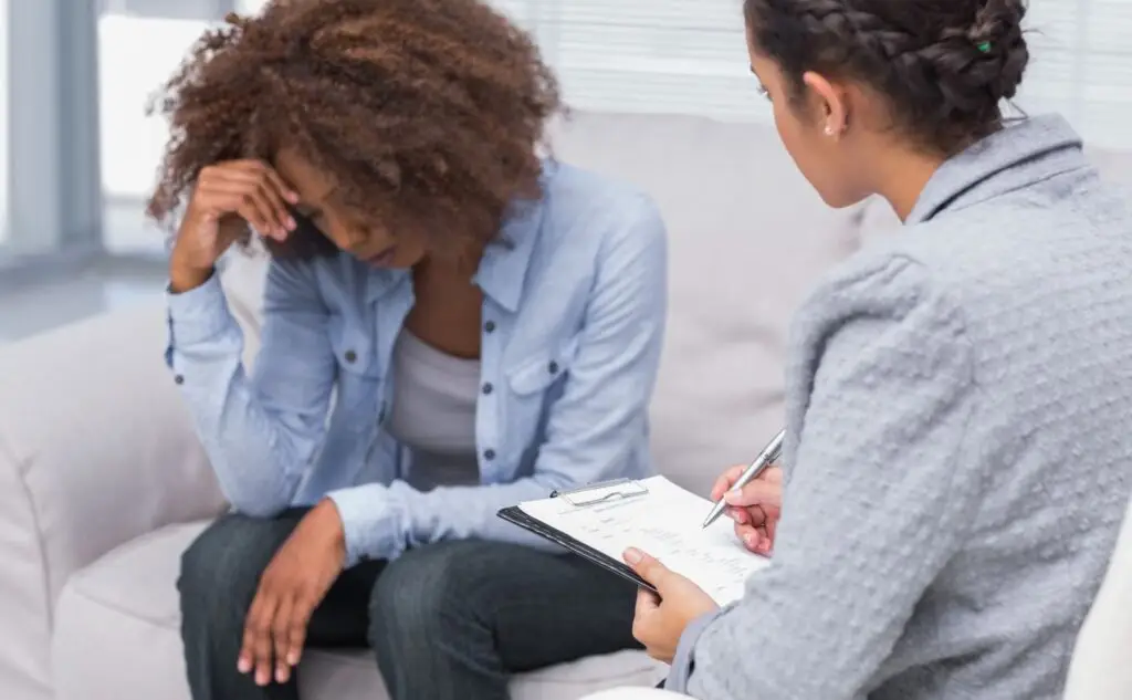 Two women in a counseling session: one looks distressed, head in hand, while the other writes notes on a clipboard nearby.