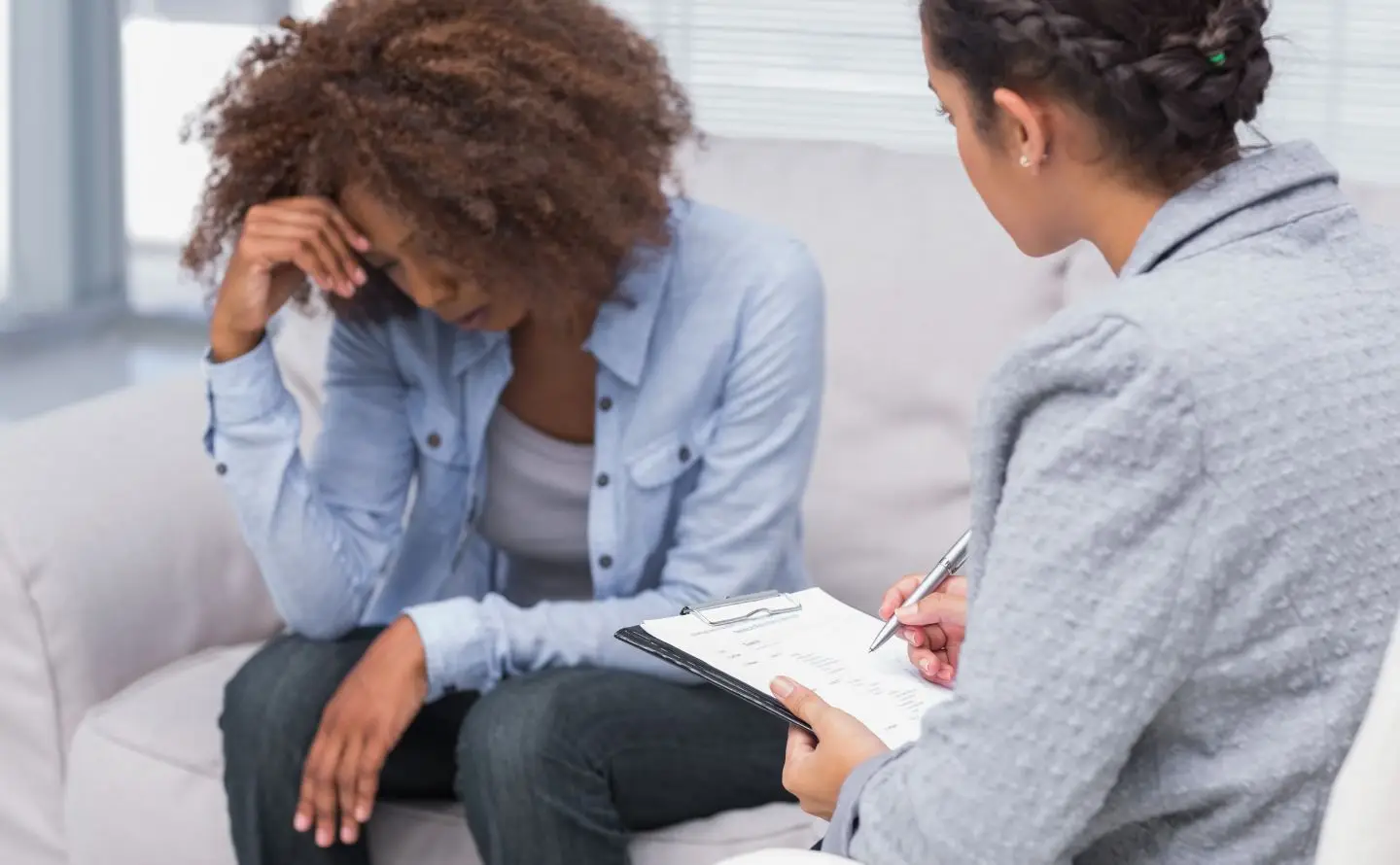 Two women in a counseling session: one looks distressed, head in hand, while the other writes notes on a clipboard nearby.