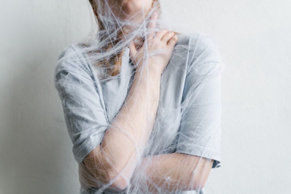 Person in a gray shirt wrapped in clear plastic wrap, arms crossed, facing a wall; conveys a sense of being trapped.