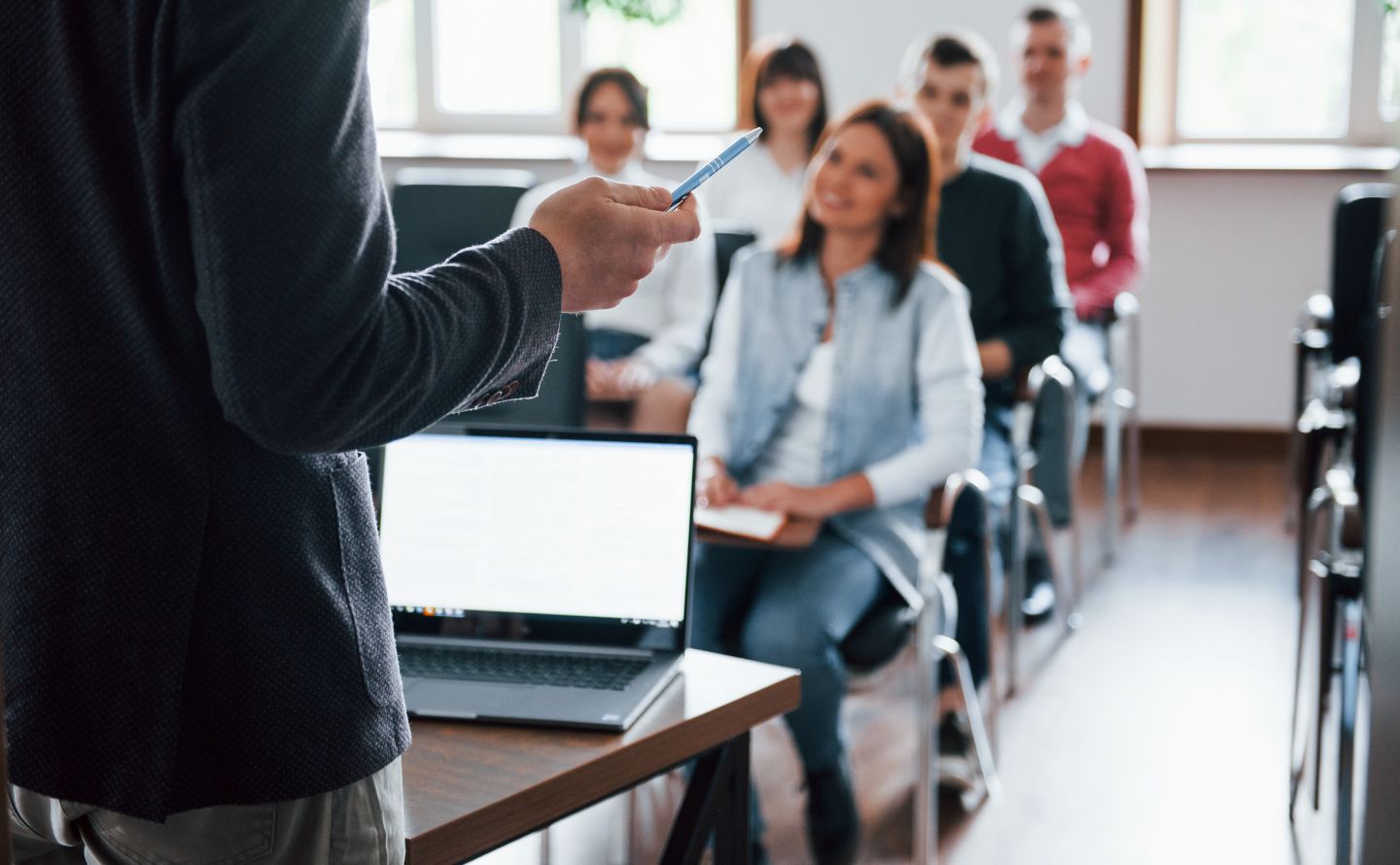 Instructor standing at a desk with a laptop, gesturing with a blue pen as a diverse group of adults sits and listens in a bright classroom seting.