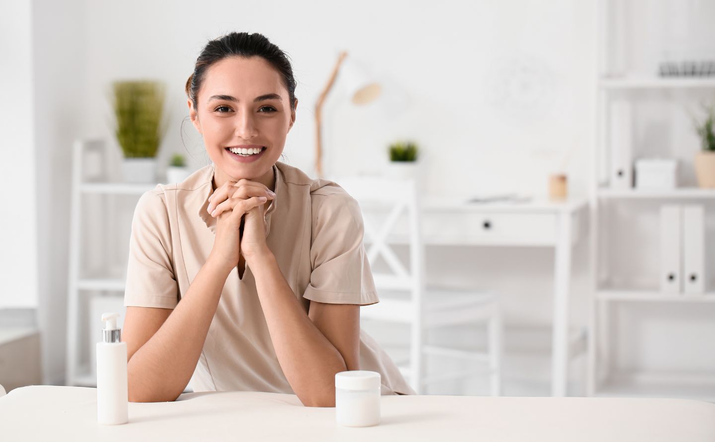 Smiling woman at a white desk with a pump bottle and a jar of cream in a bright, minimalist office.