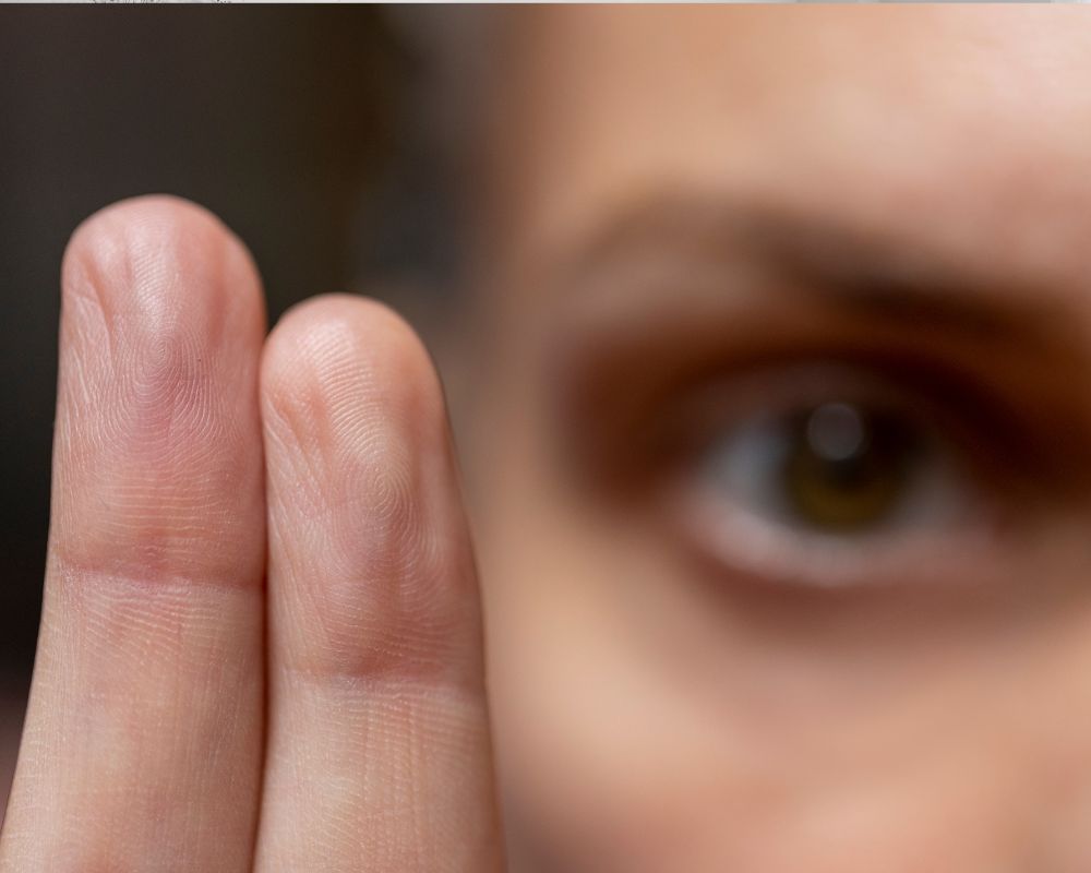 Close-up of two fingers in the foreground with a blurred eye in the background.