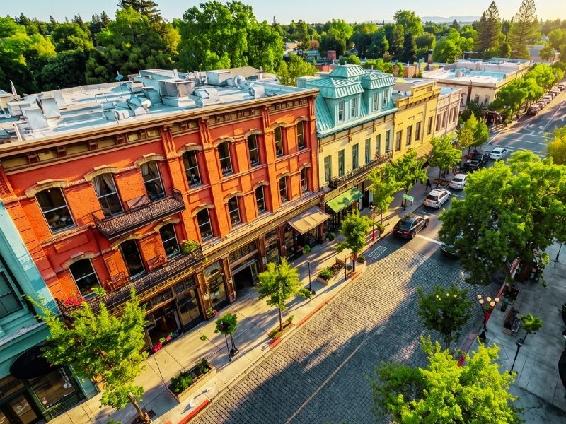 Aerial view of a sunny street with red brick storefronts, teal and yellow building facades, a cobblestone road, and tree-lined sidewalks.