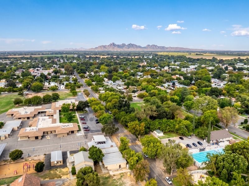 Aerial view of a small town with tree-lined streets, low-rise buildings, and distant mountains under a blue sky.