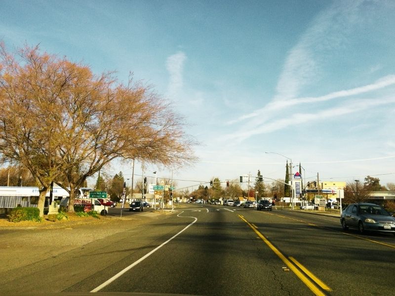 Two-lane road leads straight through a small town with leafless trees on the left, cars parked and driving, traffic lights ahead under a blue sky