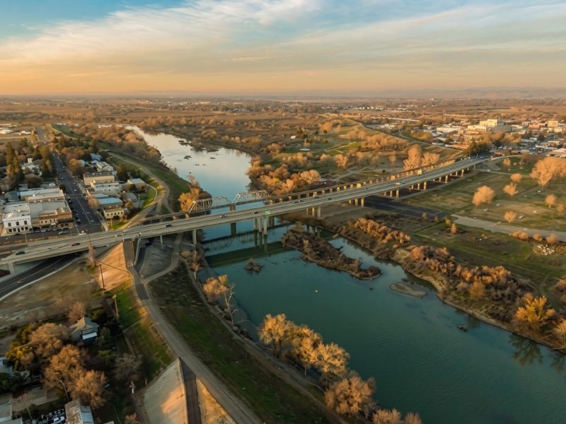 Aerial view of a river crossing through a town with parallel bridges and surrounding roads and trees at sunset.